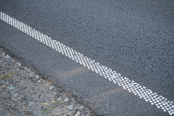 A close view of a black asphalt road with textured white dotted edge markings. The natural terrain is visible alongside the road, showing gravel and grass patches