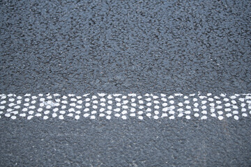 A white dotted line is painted on a black asphalt road surface, clearly marking the lanes in a busy urban environment during daylight. Vehicles can be seen in the background
