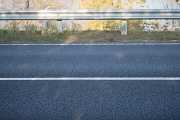 A clear view of a well-maintained asphalt road with a white striped line and a guardrail. The background features trees with leaves, indicating a sunny day