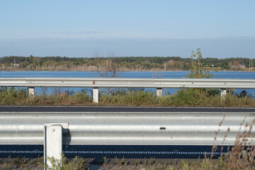 A serene view shows a lakeside environment visible from behind a metal roadside barrier. Clear skies and lush foliage create a peaceful atmosphere, reflecting nature's beauty