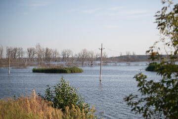 A vast body of water covers the ground, revealing patches of grass and submerged trees. Tall poles emerge from the water under a clear blue sky. It captures the aftermath of recent flooding