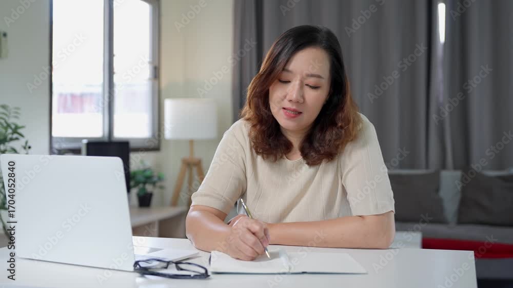 Aufkleber asian freelancer focusing intently on taking notes in notebook while working remotely from home usin