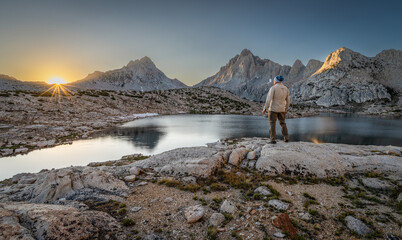 Rear view of a man standing on a rock in front of Grinnell lake at dawn looking at the Sierra Nevada Mountains, Sierra National Forest, California, USA