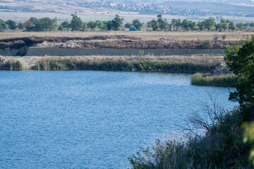 A calm lake reflects the blue sky and surrounding vegetation, with a distant town nestled beyond golden fields and trees.