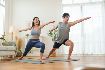 Fototapeta premium Asian couple stands strong in Warrior II pose on yoga mats, focusing together in a cozy living room.
