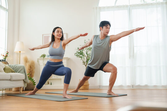 Asian couple stands strong in Warrior II pose on yoga mats, focusing together in a cozy living room.