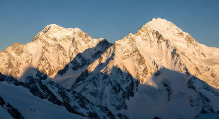 Snow capped mountain peaks bathed in golden light against a clear blue sky