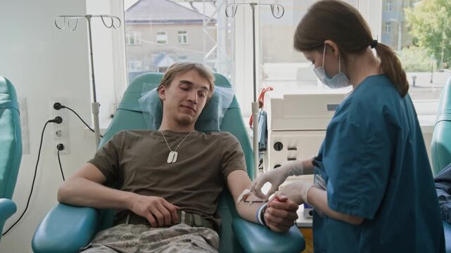 Young female nurse inserting IV line into arm of male soldier in military uniform, donating blood in clinic