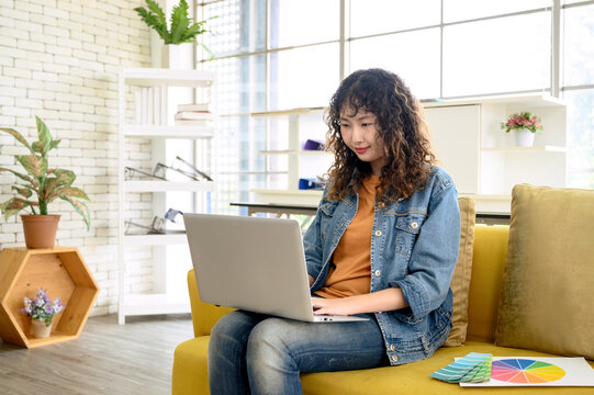Portrait of attractive young Asian female graphic designer working with laptop computer sitting on office cozy sofa.