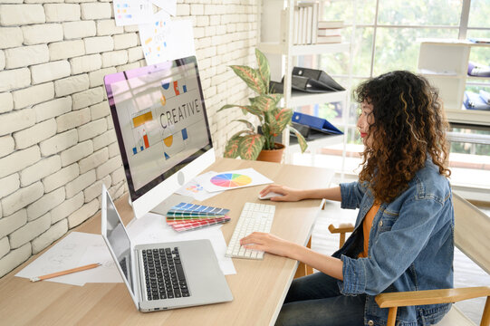 Young female graphic designers sitting at desks working with computer graphics and laptops, color charts and some sketches discussing the new project choosing a concept.