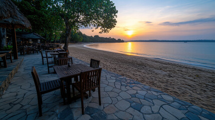 Serene Beachfront Sunset with Wooden Tables and Chairs near Calm Ocean Waves and Lush Greenery in Tranquil Environment