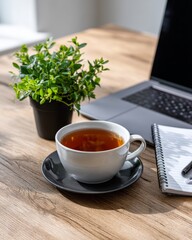 Cozy home office workspace featuring hot tea in white cup, laptop computer, green plant, and spiral notebook on natural wood desk surface