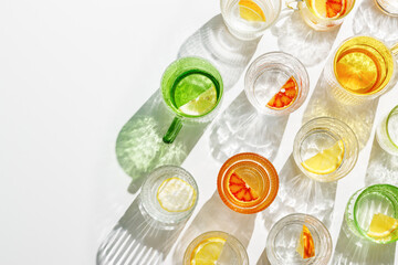 Summer still life with colored glass cups of infused water with slices of fruit on white table. Glittering different glasses at sunlight. Long harsh shadow. Summer vacation concept, top view