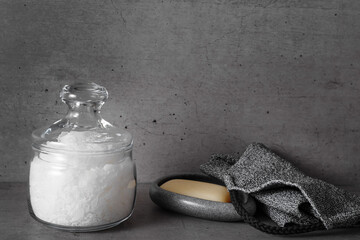 Glass jar with natural sea salt in grey bathroom, stone soap dish with soap, exfoliating body washcloth. Modern eco-friendly approach to self-care and wellness routines, home spa routine