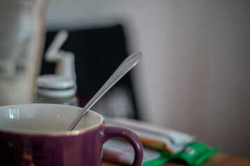 Close-up of a purple coffee mug with a metal spoon inside, placed on a messy tabletop with packages and bottles in background.