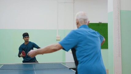 Back view of senior man in blue playing table tennis with young Indian boy across net inside indoor hall showing intergenerational training teamwork focus competition