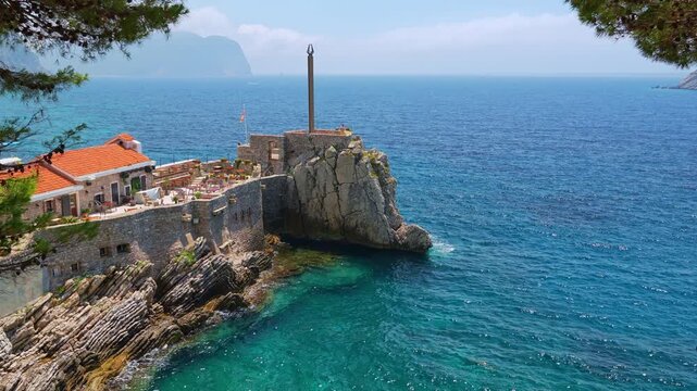 beautiful view of open-air cafe or restaurant on top of rocky coast, Adriatic Sea and bay are visible through tree branches