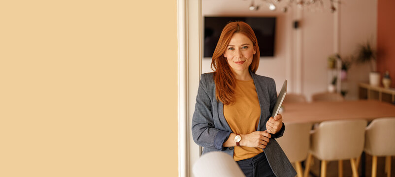 Portrait of confident female professional holding laptop and smiling at camera while leaning on wall in office