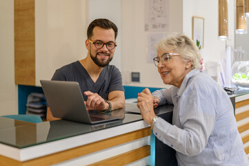 Male doctor showing laptop to smiling senior woman at reception counter in hospital during visit for eye exam