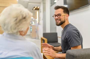 Smiling male doctor in eyeglasses explaining to senior patient while sitting together in hospital lobby