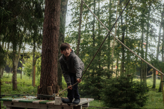 A little boy in a gray hoodie balances carefully on a tightrope walk platform, holding onto ropes at a wooded outdoor adventure park. 