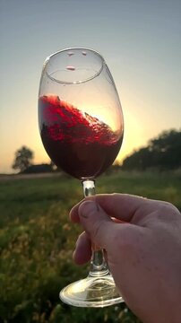 Red wine swirling in hand. Sommelier swirls with glass of red wine at sunset. Red Wine Swirling In Glass during vine Tasting. Celebration vino party in countryside. Glass in hand on Rural landscape. 