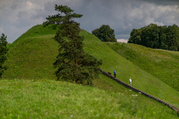 Scenic landscape with green grassy hills and a wooden staircase leading up the slope. A few people are walking along the stairs, surrounded by lush nature and dramatic cloudy sky. 