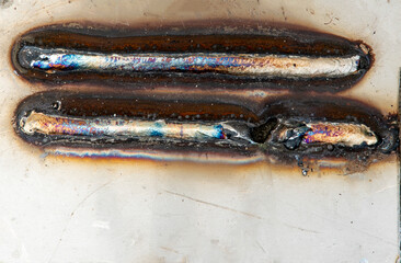 Close-up of several welding test beads on metal plates, showing different welding techniques and heat discoloration. Industrial background with practice welds on steel sheets placed on the ground."