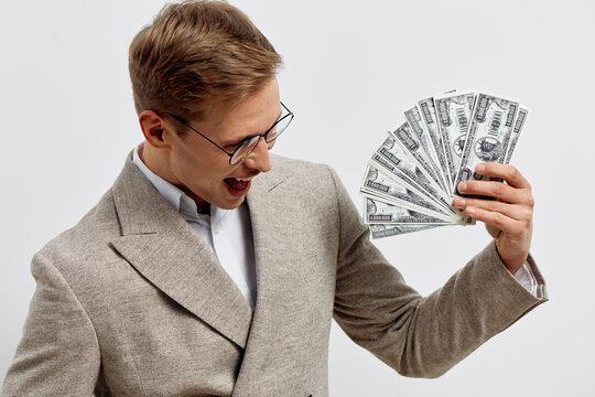 Confident young man with glasses wearing beige blazer holds fan of hundred dollar bills joyful expressively isolated on plain white background studio portrait. Finance success wealth concept. - Powered by Adobe