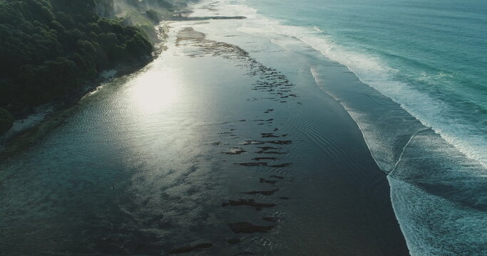 Aerial view capturing sunlight shimmering on shallow tropical waters, accompanied by gentle incoming waves and lush green cliffs under a bright morning sky - Powered by Adobe