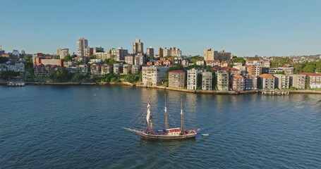 Australia, Sydney: Aerial drone revealing Sydney's skyline with a traditional sailing ship navigating the harbor, blending modern architecture and maritime heritage. Aerial view drone