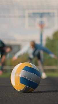 Close-up of volleyball resting on outdoor court with blurred view of athletes performing stretches and bends in the background, sports facility, and building visible