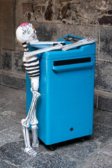 A skeleton is sitting on top of a blue box. The skeleton is decorated with black and white stripes. Day of the Dead celebration in Mexico, papel picado crafts, offerings, La Catrina