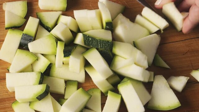 A woman's hand slices green zucchini on a cutting board on the table with a knife. Medium shot.