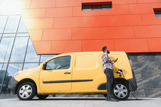 Delivery driver charging electric van and using smartphone
