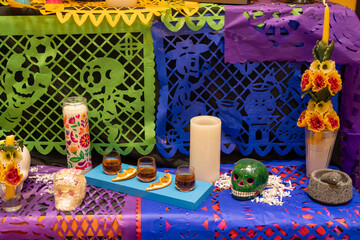 A table with a Mexican themed display of candles, skulls, and flowers. The table is covered with colorful paper. Day of the Dead celebration in Mexico, papel picado crafts, offerings, La Catrina