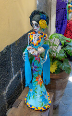 Fototapeta premium A skeleton dressed in a blue dress with flowers and a flower crown. The skeleton is holding a cup in one hand. Day of the Dead celebration in Mexico, papel picado crafts, offerings, La Catrina