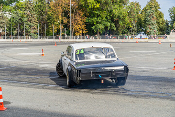 Racing car drifting on an asphalt track between cones. Tire smoke and spectators behind barriers create a competitive atmosphere.