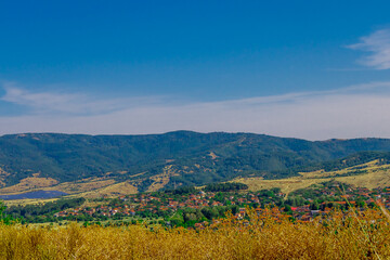 Obraz premium Picturesque rural village at the foot of green mountains with golden summer fields under a bright blue sky