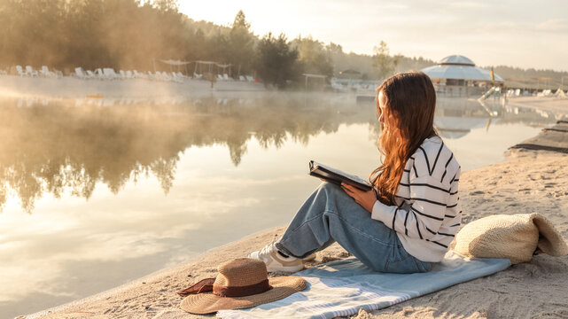 Young woman reading a book by the lake at sunrise