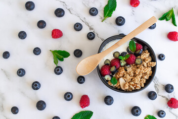 Top view of a yogurt bowl with granola, raspberries, blueberries, and mint on a white marble surface.