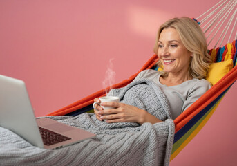 Smiling mature woman relaxing in a cozy hammock with a laptop and coffee. Lifestyle and remote work concept. Copy space on a pink background