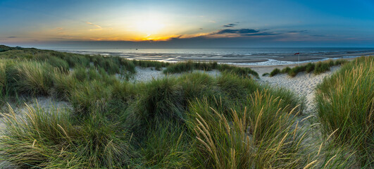 North Sea beach with reed grass bushes at sunset, orange-red sky over the dark blue ocean, panorama with sandy beach and blowing grass in the wind, sunset over a wall of clouds, fisher boat on tour