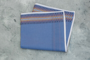 A neatly folded men's handkerchief with a blue base and a patterned band of woven stripes rests on a textured gray background.