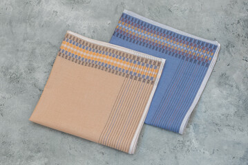 Two neatly folded men's handkerchiefs, one in blue and one in beige, both with a woven geometric pattern, are displayed side by side on a textured gray surface.