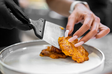 Close-up of hands plating crispy fried food with spatula onto white dish. Black glove and manicured nails highlight contrast in kitchen setting. Culinary preparation concept.