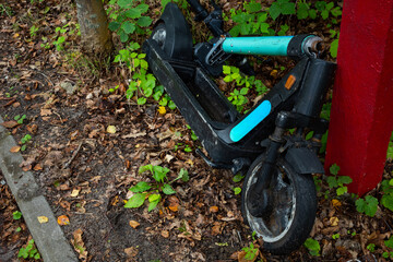 A damaged public electric scooter lies on the side of the road. The photo was taken on a cloudy day with natural lighting.