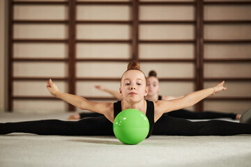 Portrait of Caucasian girl gymnast performing split stretch on floor with arms extended, focusing on green ball in foreground, another girl gymnast visible in background