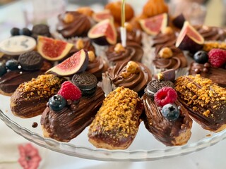 Chocolate eclairs topped with figs, berries, cookies, and nuts served on a glass cake stand.