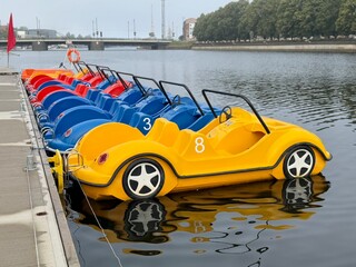 Row of colorful pedal boats shaped like cars docked on calm water, ready for rental and leisure rides.